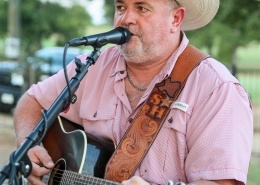 A man wearing a cowboy hat plays acoustic guitar and sings into a microphone during an outdoor performance.